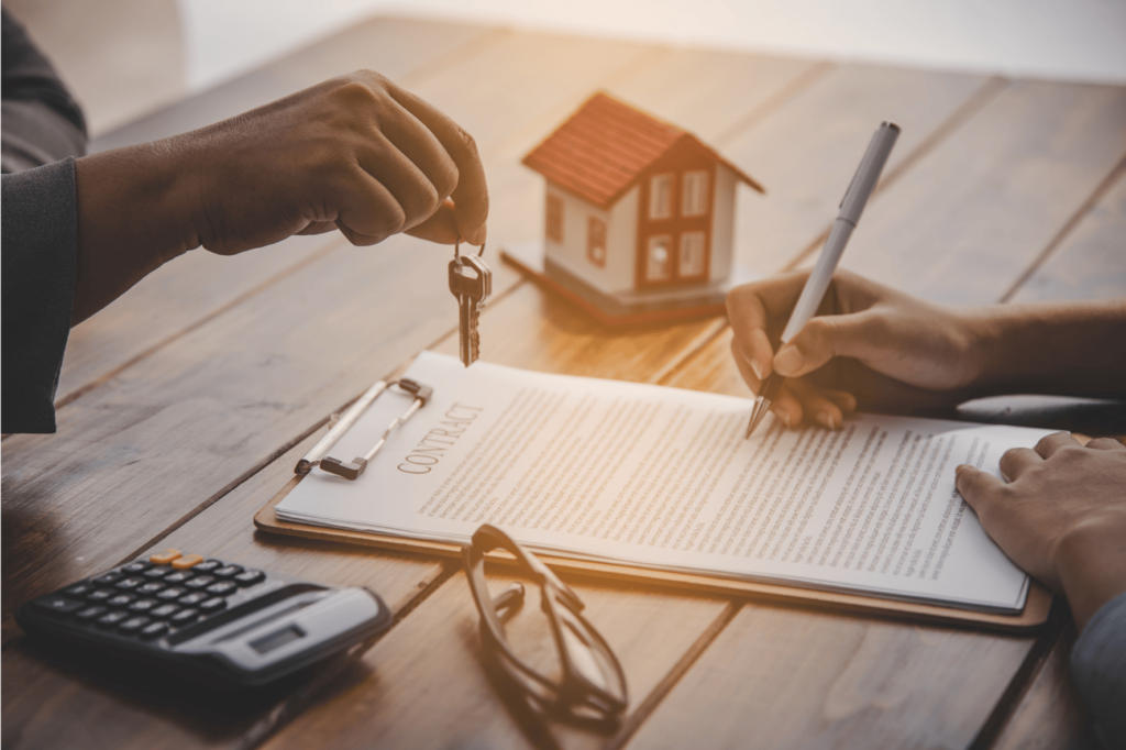 Les mains d'une personne sont visibles sur une table en bois. Une main, à gauche, tient un trousseau de clés. L'autre main, à droite, écrit sur un contrat de prêt immobilier posé sur un presse-papiers. À l'arrière-plan, à gauche de la main qui écrit, il y a une calculatrice, et à sa droite, une paire de lunettes. Derrière le contrat, une petite maison miniature est posée sur la table. La scène est bien éclairée depuis le haut à gauche, diffusant une lueur chaleureuse.