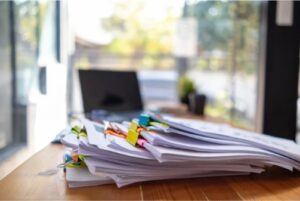 Une pile de documents et de papiers, certains attachés par des trombones de différentes couleurs, est posée sur un bureau en bois. À l'arrière-plan flou, on aperçoit un ordinateur portable et une fenêtre donnant sur l'extérieur. Cette image peut symboliser les documents nécessaires ou demandés lors de la souscription à un prêt personnel, les justificatifs à fournir pour obtenir ce type de financement.