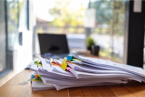 Une pile de documents et de papiers, certains attachés par des trombones de différentes couleurs, est posée sur un bureau en bois. À l'arrière-plan flou, on aperçoit un ordinateur portable et une fenêtre donnant sur l'extérieur. Cette image peut symboliser les documents nécessaires ou demandés lors de la souscription à un prêt personnel, les justificatifs à fournir pour obtenir ce type de financement.