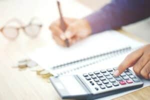 L'image montre une personne utilisant une calculatrice avec sa main droite, tandis que sa main gauche tient un stylo au-dessus d'un carnet à spirale ouvert. Des pièces de monnaie sont éparpillées sur le bureau en bois à côté de lunettes, créant une atmosphère de calcul financier ou de budgétisation. La mise au point est sur les mains et les objets au premier plan, avec un arrière-plan légèrement flou et lumineux.