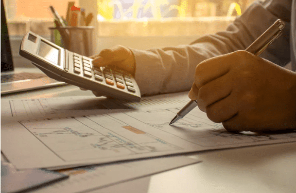 Une personne est assise à un bureau, ses mains occupées. La main gauche utilise une calculatrice, tandis que la main droite tient un stylo et est posée sur des documents financiers, possiblement liés à un rachat de crédit ou à la gestion budgétaire. À l'arrière-plan, un ordinateur portable est partiellement visible sur la gauche, et un pot à crayons se trouve sur la droite, avec une lumière diffuse provenant d'une fenêtre. L'accent est mis sur les mains et les outils de travail.