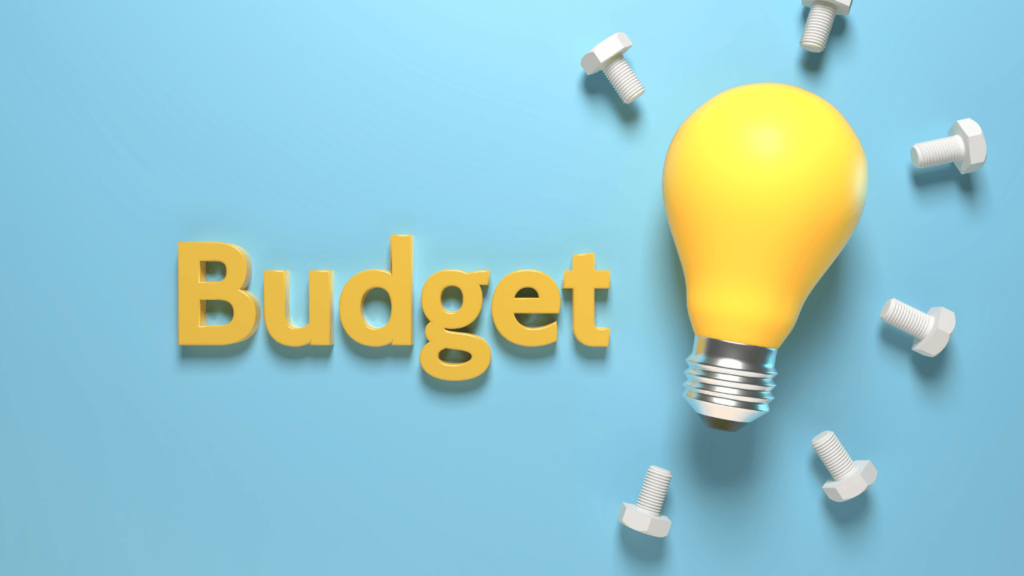 Le mot "Budget" en 3D de couleur jaune est posé sur une surface bleue. À côté, une ampoule jaune et lumineuse est entourée de plusieurs boulons et écrous blancs dispersés. L'image pourrait symboliser des idées ou des solutions pour la gestion du budget, potentiellement en lien avec un rachat de crédit, où l'optimisation des dépenses est essentielle.
