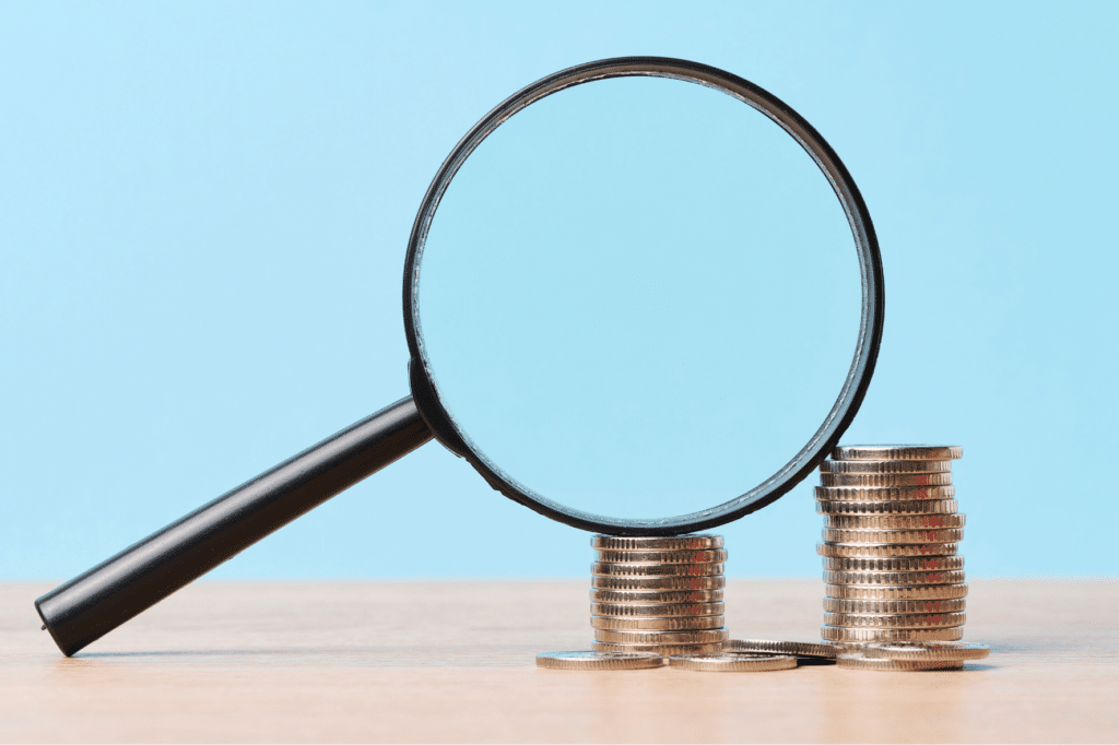 Une loupe repose contre deux piles de pièces de monnaie, avec quelques pièces supplémentaires éparpillées à la base des piles, le tout sur une surface en bois avec un arrière-plan bleu clair.