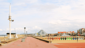 L'image présente une vue de la plage et de la promenade du Touquet-Paris-Plage, sous un ciel parsemé de nuages. Au premier plan à droite, une allée pavée de couleur rouille s'étend le long de la plage. Sur la gauche, on voit un large banc de sable. Des cabines de plage colorées (blanches, jaunes, rouges, vertes, bleues) bordent le sable le long de la promenade. Au loin, le bâtiment pyramidal de l'Aqualud est visible, ainsi que d'autres constructions caractéristiques de la ville du Touquet avec leurs toits rouges.