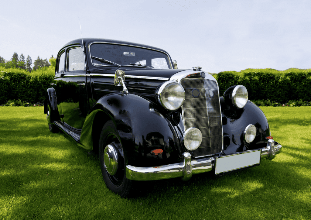 Une berline de collection Mercedes-Benz 170 S noire est garée sur l'herbe d'un parc lors d'une journée ensoleillée. La voiture a un design classique avec une carrosserie élégante, des phares ronds et une calandre chromée.