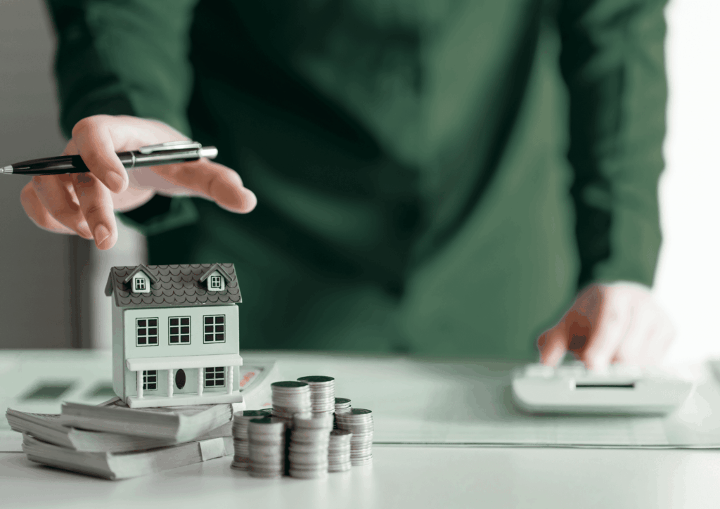 Maquette de maison, pièces et liasses de billets posées sur une table, avec une personne tenant un stylo et une calculatrice, illustrant les liquidités que peut dégager une vente à réméré ainsi que les couts associés comme les indemnités d'occupation