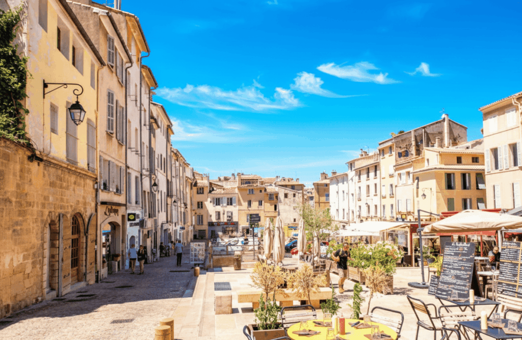 Rue piétonne ensoleillée dans la vieille ville d'Aix-en-Provence (ou du sud de la France), avec des façades en stuc jaune et des cafés en terrasse. Une table de café jaune est visible au premier plan sur une place pavée.