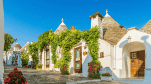 Un plan extérieur d'une rue pavée bordée de 'trulli', les maisons traditionnelles blanchies à la chaux avec des toits coniques en pierre d'Alberobello, dans les Pouilles, en Italie. Les maisons sont ornées de feuillage vert grimpant et de fleurs roses et rouges vives. Le ciel est d'un bleu clair intense sans nuages.