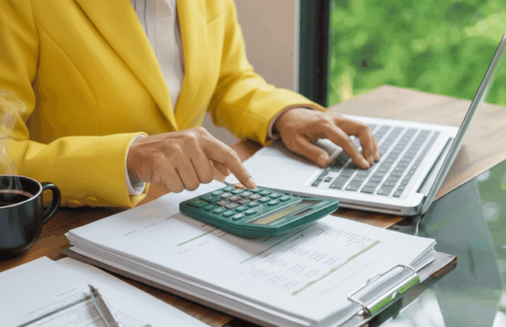 Gros plan sur une personne en veste jaune travaillant sur un ordinateur portable et utilisant une calculatrice pour payer une dette fiscale, avec une tasse de café à côté de dossiers professionnels