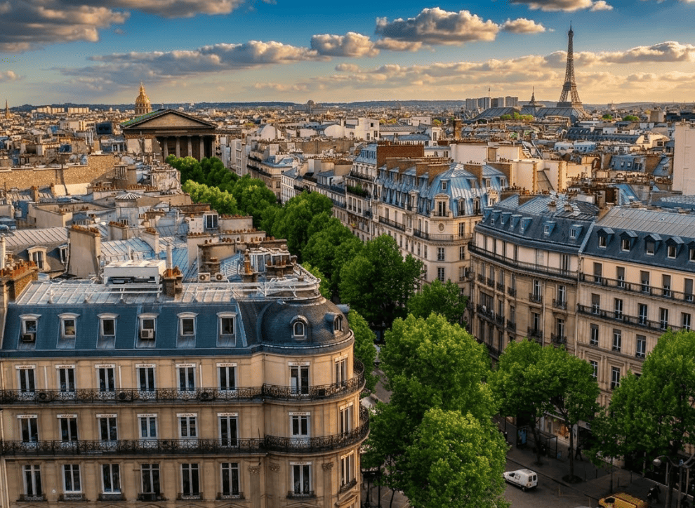 Une vue panoramique en plongée des toits de Paris sous un ciel de fin de journée ensoleillé. Au premier plan, des immeubles haussmanniens classiques avec des balcons en fer forgé et des toits en zinc gris. Une large avenue bordée d'arbres verdoyants mène vers l'église de la Madeleine sur la gauche, tandis que la Tour Eiffel se dresse fièrement à l'horizon sur la droite. La lumière chaude du soleil couchant illumine les façades en pierre calcaire, contrastant avec un ciel bleu parsemé de quelques nuages cotonneux.