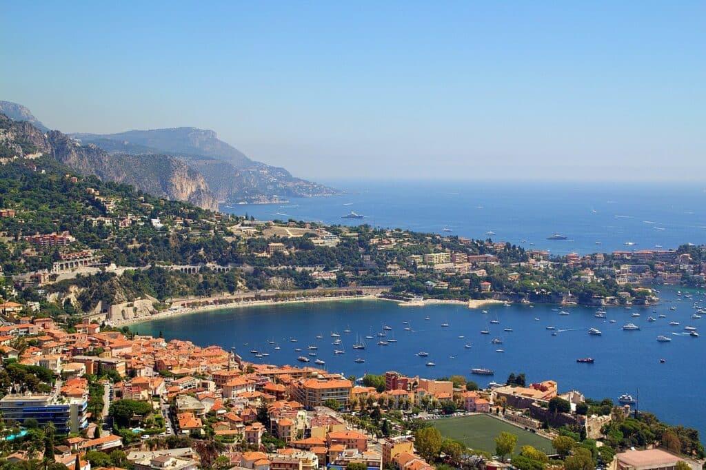 Vue panoramique en plongée d'une baie sous un ciel bleu dégagé. Au premier plan, les toits de tuiles orange de la vieille ville et un terrain de sport verdoyant. Dans la baie azur, de nombreux bateaux et yachts sont à l'ancre. À l'arrière-plan, les collines escarpées de la Côte d'Azur s'étendent vers l'horizon. Environnement idéal pour l'achat d'une résidence secondaire